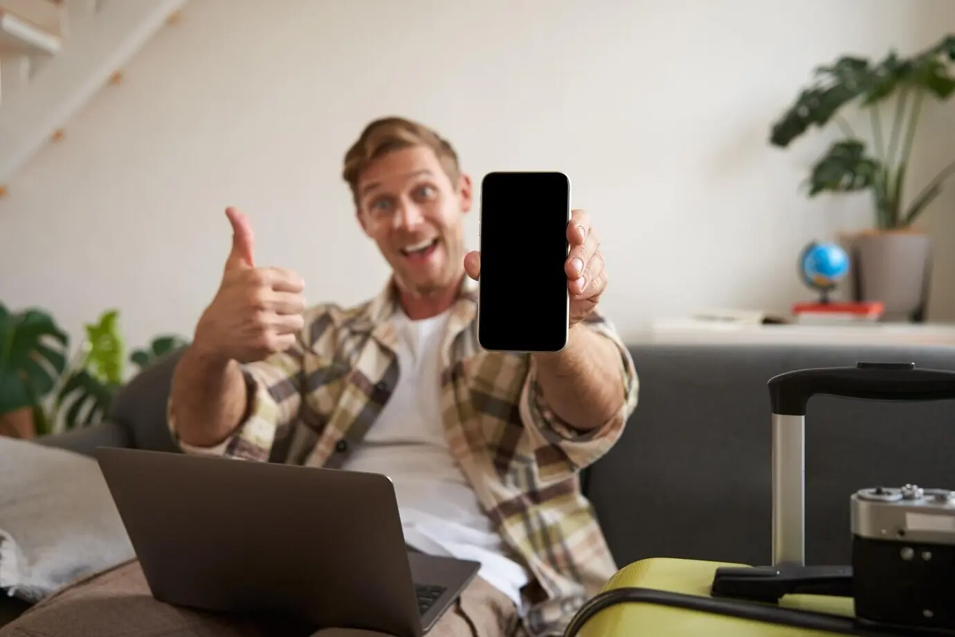 Portrait of a male tourist showing his mobile phone screen, recommending a travel app, while sitting with a suitcase.