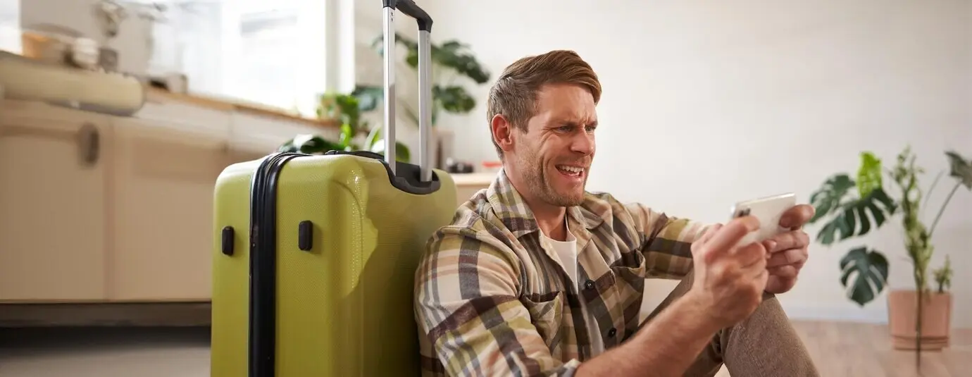 A man with a shocked expression sits on the floor with a suitcase, looking worried and tense at his mobile phone screen.