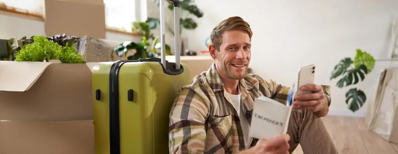 A portrait of a handsome, smiling male tourist holding a passport and flight tickets, seated with a suitcase.