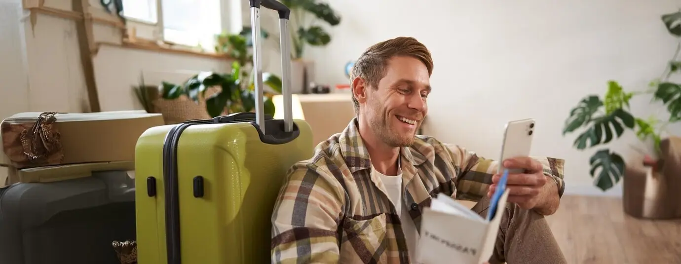 Portrait of a smiling male tourist sitting with a suitcase, holding a smartphone, passport, and tickets, going on holiday.